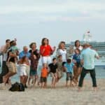 A group of people, including adults and children, are jumping on a beach while being photographed, with a large cruise ship in the background.