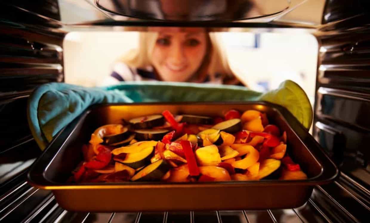 A woman is looking into an oven where a tray of chopped vegetables is roasting, using the leftover heat to ensure a perfect cook. She appears pleased and is wearing a blue and white striped shirt. The tray is lined with oven mitts for handling.