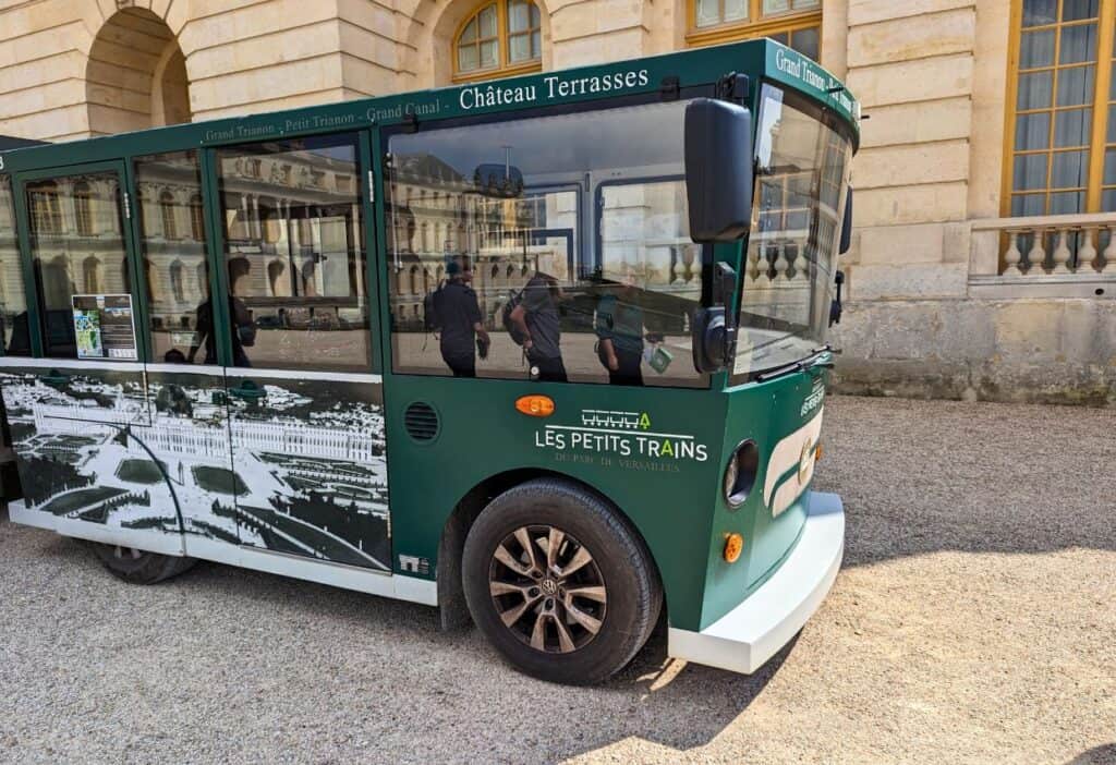 A green and white tour tram labeled "Les Petits Trains" is parked in front of a historical building with arched windows. Three people are visible through the tram windows, conversing.