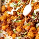 A close-up of loaded tater tots topped with melted cheese, bacon, chopped green onions, and a drizzle of creamy sauce being added from a white spoon.