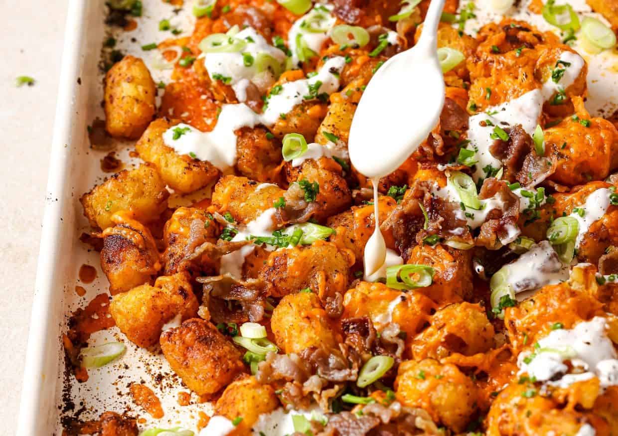 A close-up of loaded tater tots topped with melted cheese, bacon, chopped green onions, and a drizzle of creamy sauce being added from a white spoon.
