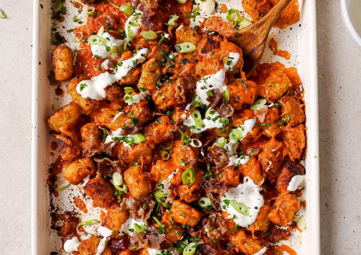 A close-up of loaded tater tots topped with melted cheese, bacon, chopped green onions, and a drizzle of creamy sauce being added from a white spoon.