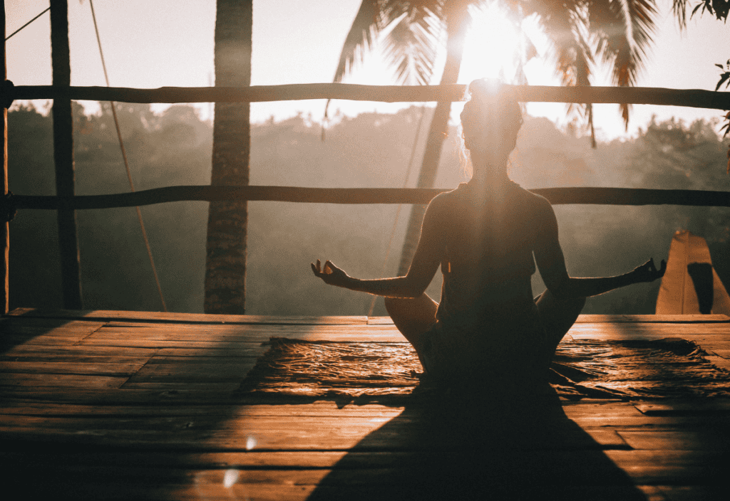 A person sits in a lotus position on a mat, meditating on a wooden deck at sunrise. Palm trees and sunlight fill the background.