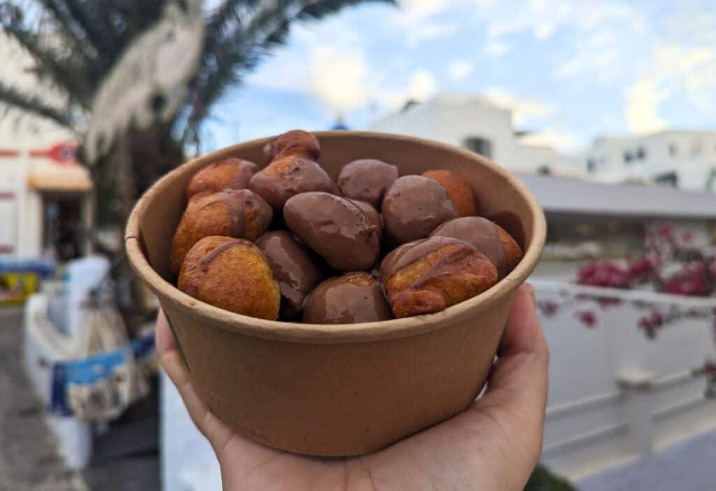 A hand holds a brown paper bowl filled with small round pastries coated in chocolate, with a scenic outdoor background.