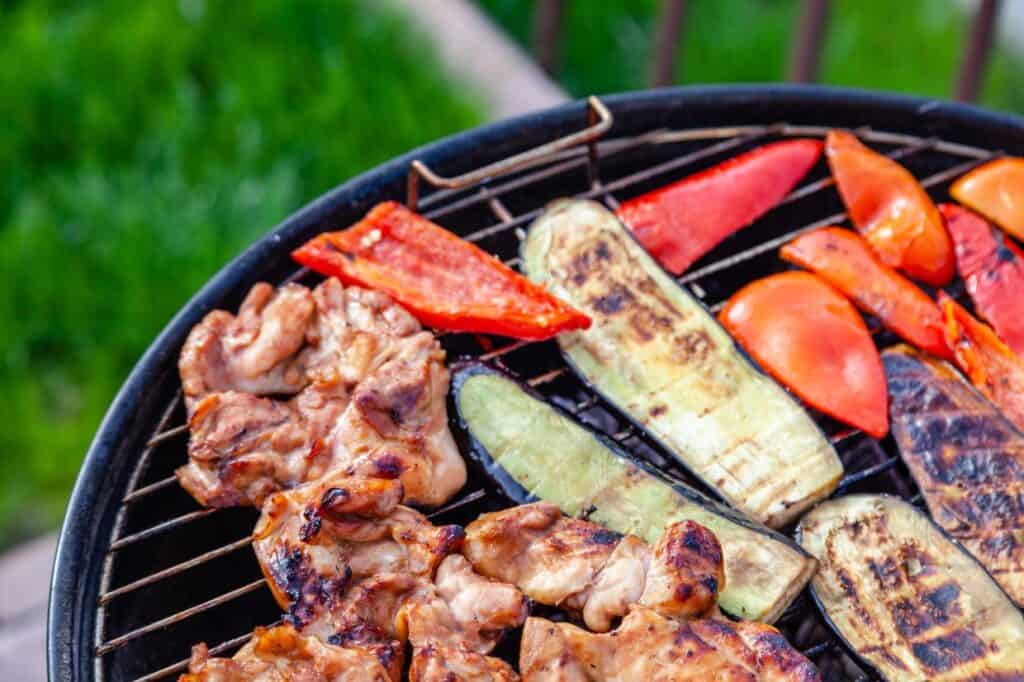 A grill with assorted foods including chicken pieces, red bell pepper slices, and zucchini slices being cooked outdoors.