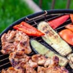 A grill with assorted foods including chicken pieces, red bell pepper slices, and zucchini slices being cooked outdoors.