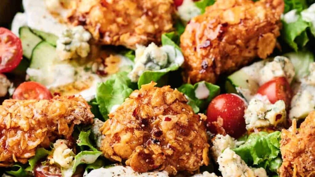 Close-up of a salad with crispy chicken pieces, cherry tomatoes, cucumbers, blue cheese crumbles, and lettuce, dressed with a light sauce.