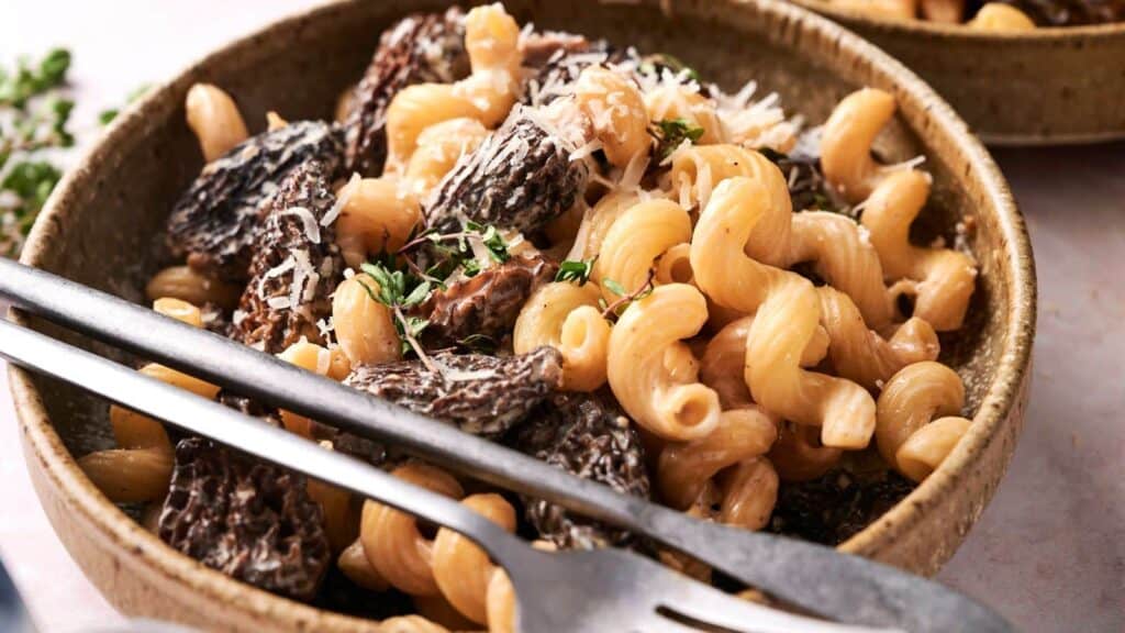 A bowl of cavatappi pasta with morel mushrooms, garnished with grated cheese and herbs, with a fork and spoon placed beside it.