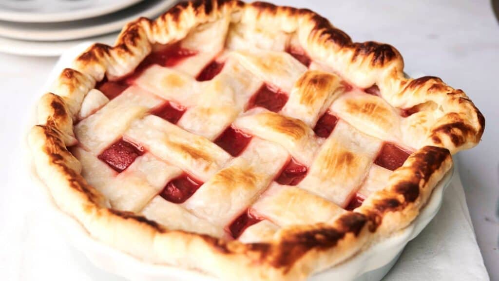 A freshly baked pie with a lattice crust, showing a golden-brown top and red filling, sits in a white pie dish.
