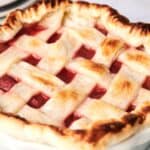 A freshly baked pie with a lattice crust, showing a golden-brown top and red filling, sits in a white pie dish.