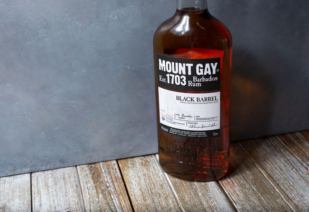 A bottle of Mount Gay Black Barrel rum sits on a wooden surface against a gray background.