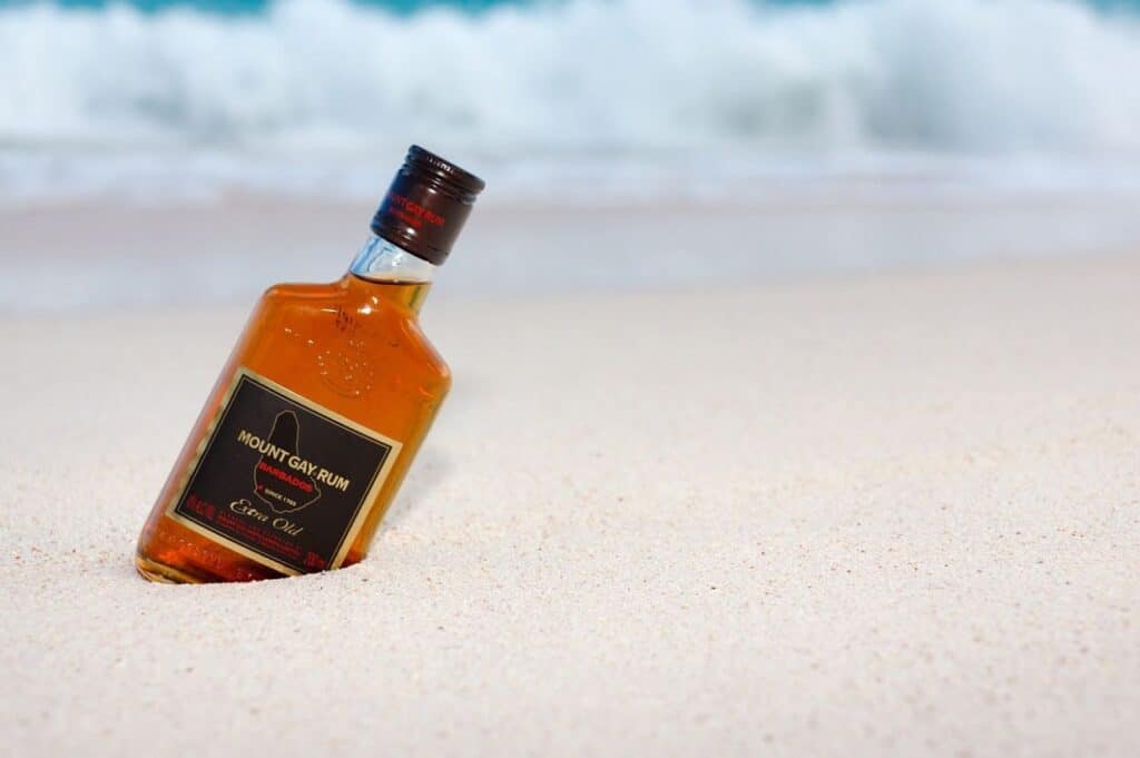 A bottle of Mount Gay Rum is partially buried in the sand on a beach with waves in the background.