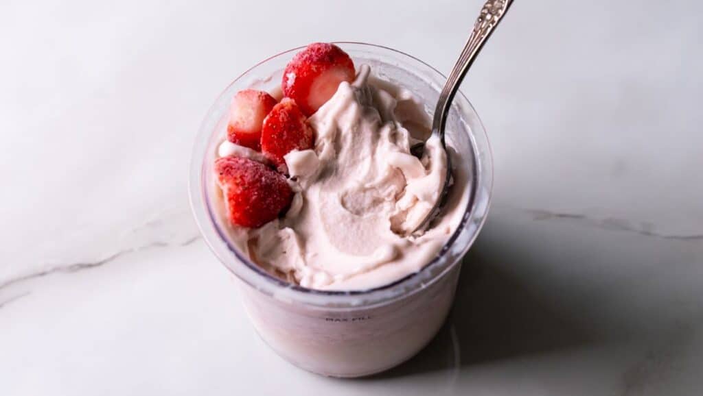 A cup of strawberry yogurt topped with fresh strawberry slices and a silver spoon on a white marble surface.
