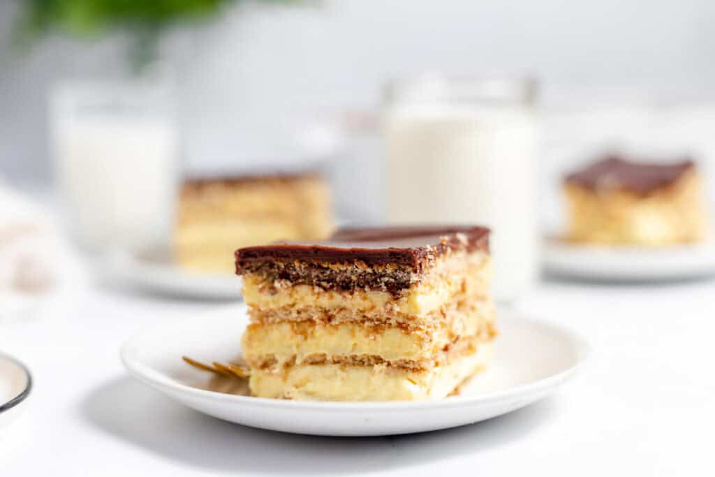 A slice of layered dessert with chocolate topping and creamy filling on a white plate, with two glasses of milk and more dessert slices blurred in the background.