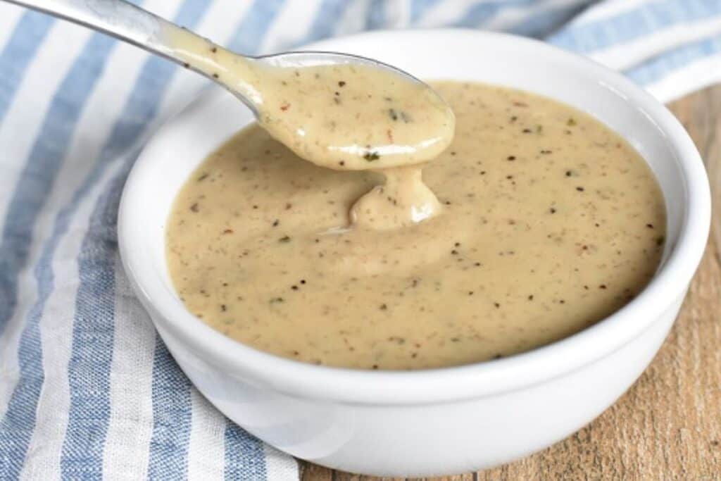 A spoon dipping into a bowl of creamy peppercorn gravy placed on a striped blue and white cloth.