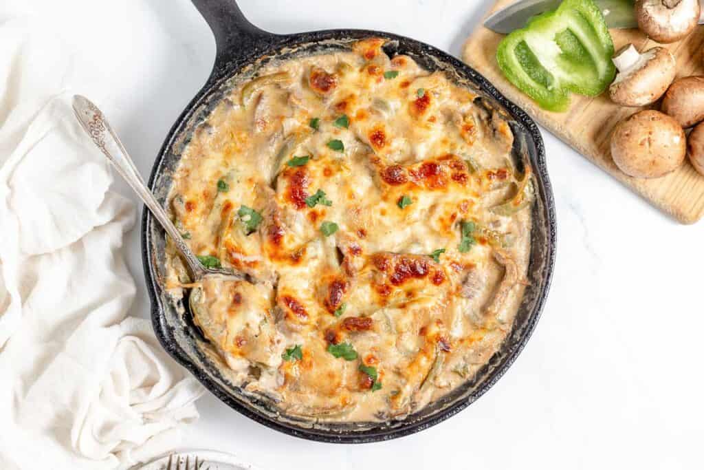A skillet containing a creamy casserole topped with melted cheese, garnished with herbs, placed on a white surface next to a chopping board with sliced green bell pepper and mushrooms.