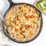 A skillet containing a creamy casserole topped with melted cheese, garnished with herbs, placed on a white surface next to a chopping board with sliced green bell pepper and mushrooms.