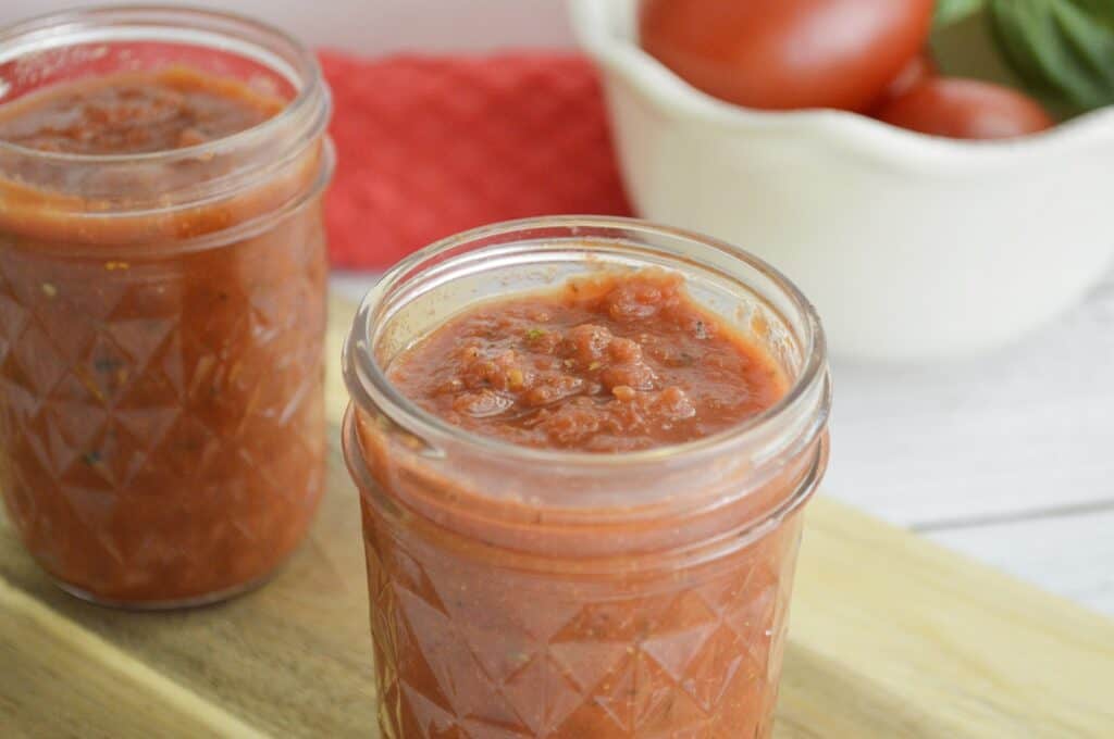 Two jars of tomato sauce are placed on a wooden surface. In the background, there are tomatoes and basil leaves in a white bowl.