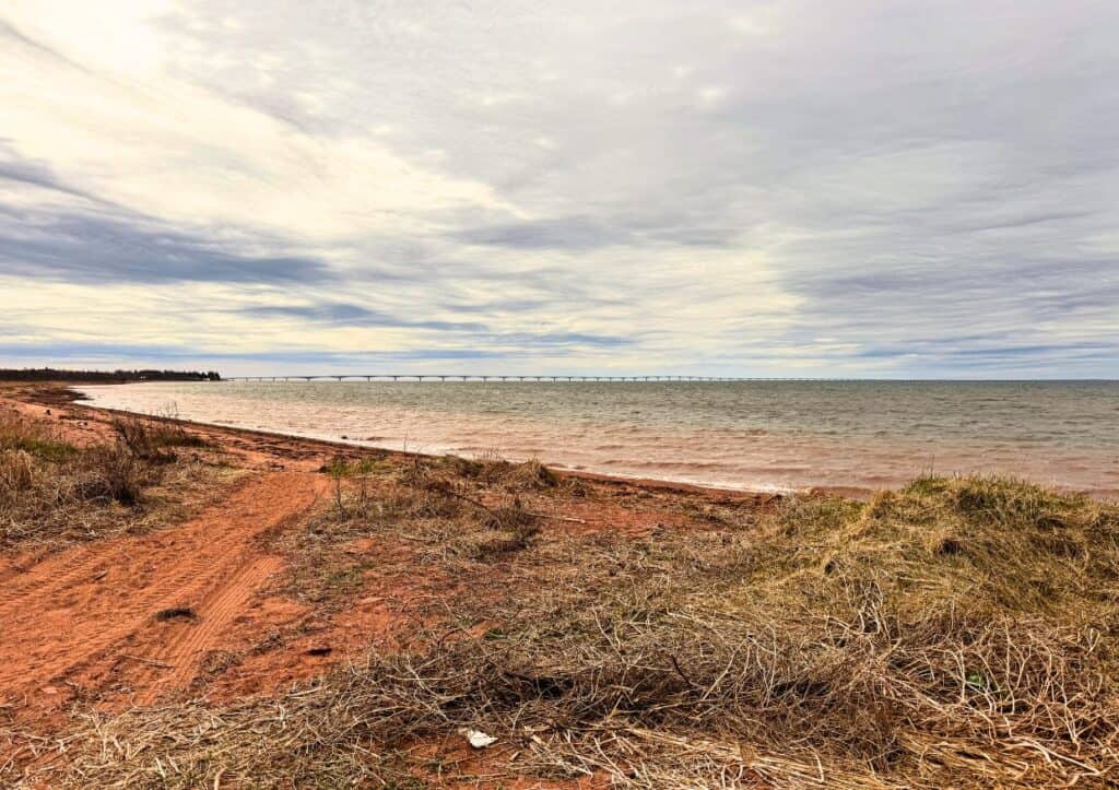 A coastline with reddish sand and grassy patches leads to a large bridge in the distance under a cloudy sky.