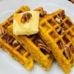 A plate of three pumpkin waffles topped with a pat of butter, syrup, and pecans.