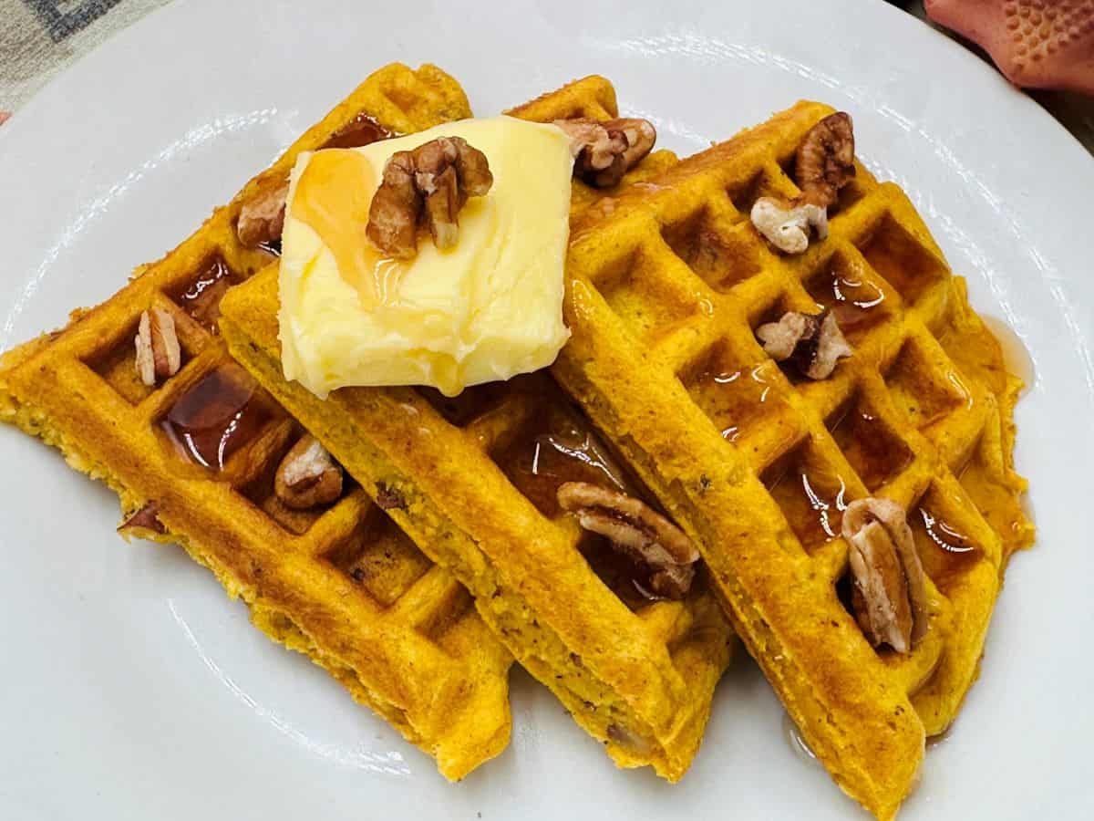 A plate of three pumpkin waffles topped with a pat of butter, syrup, and pecans.