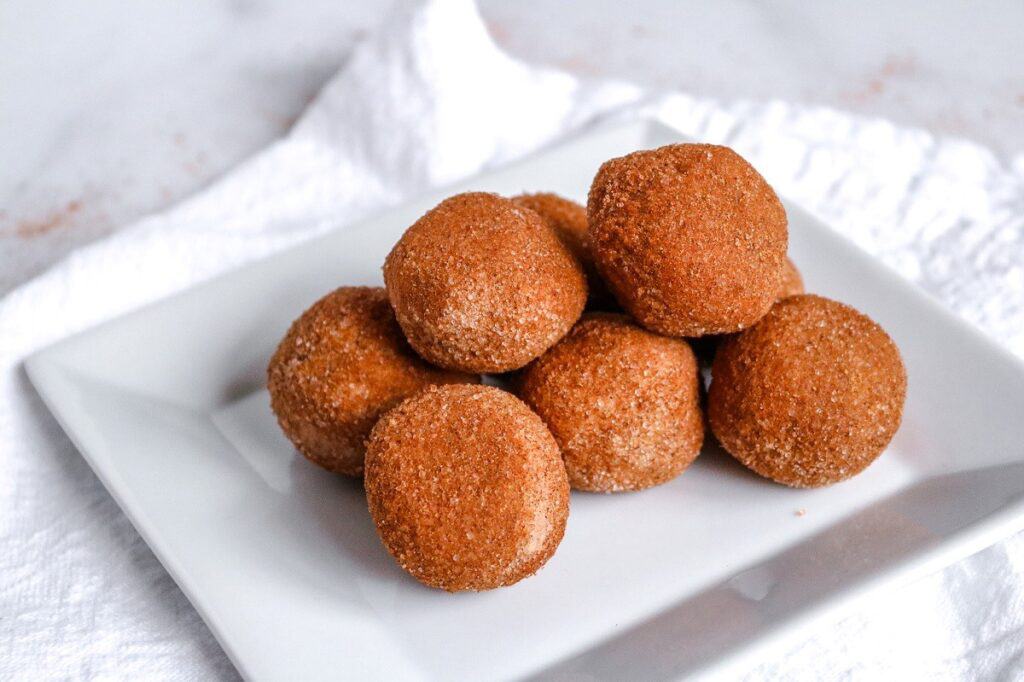 A square white plate with a pile of small, round, and browned dough balls, possibly covered in a sugar and spice mixture, placed on a white cloth background.