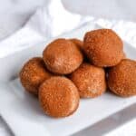 A square white plate with a pile of small, round, and browned dough balls, possibly covered in a sugar and spice mixture, placed on a white cloth background.