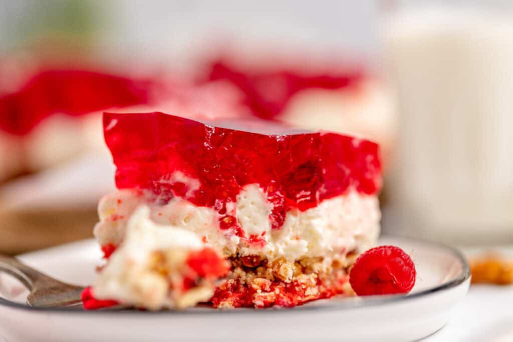 A close-up of a dessert slice with layers of red gelatin, creamy filling, and a crumbly crust on a white plate, accompanied by a fresh raspberry and a blurred background.