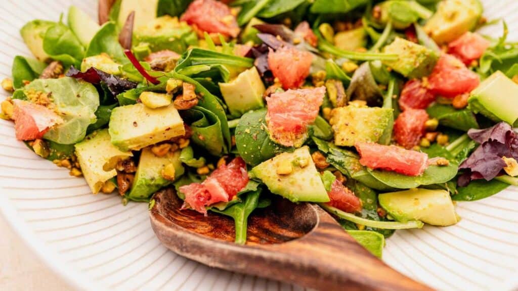 A fresh salad consisting of mixed greens, avocado chunks, grapefruit segments, and pistachios sits in a white dish, with a wooden serving spoon placed at the bottom of the dish.