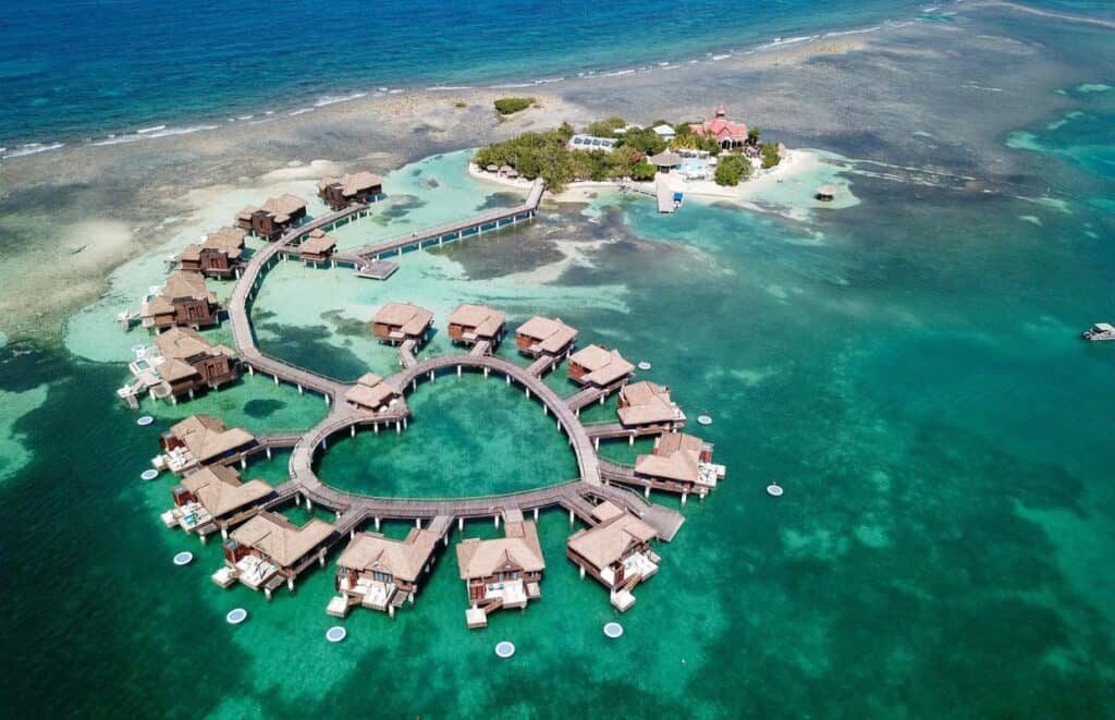 Aerial view of overwater bungalows arranged in a heart shape, surrounded by turquoise ocean water, with a small island and additional structures in the background—perfect for solo travelers seeking serenity at Caribbean resorts.