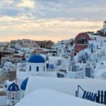 A picturesque view of Santorini, Greece, featuring a cluster of white buildings with blue domes against a backdrop of a cloudy sky.