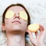 Person lying down with potato slices on their eyes, holding another slice near their face, on a fluffy surface.