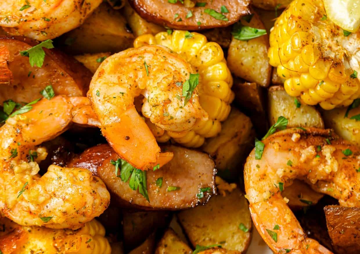 A dish of seasoned shrimp, corn on the cob, sausage slices, and potato chunks garnished with fresh herbs and lemon wedges.