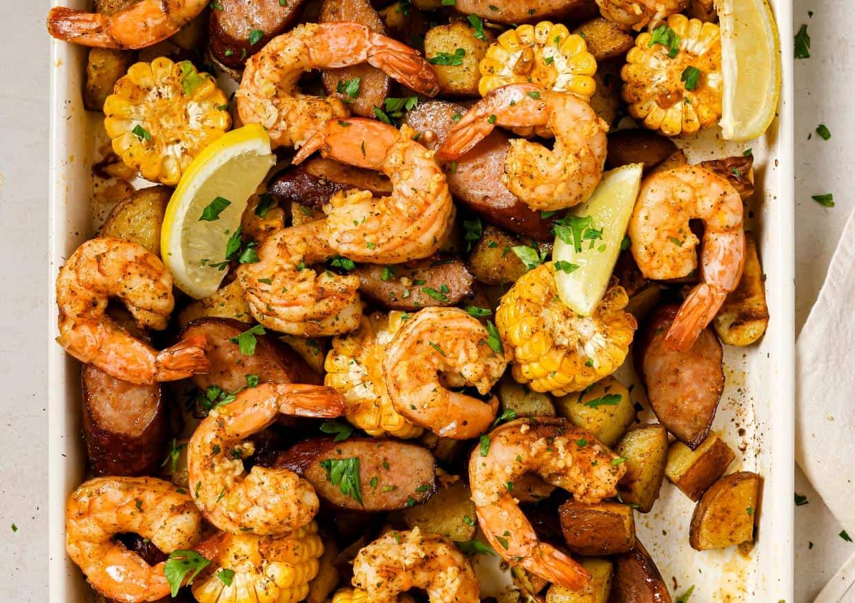 A dish of seasoned shrimp, corn on the cob, sausage slices, and potato chunks garnished with fresh herbs and lemon wedges.