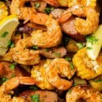 A dish of seasoned shrimp, corn on the cob, sausage slices, and potato chunks garnished with fresh herbs and lemon wedges.