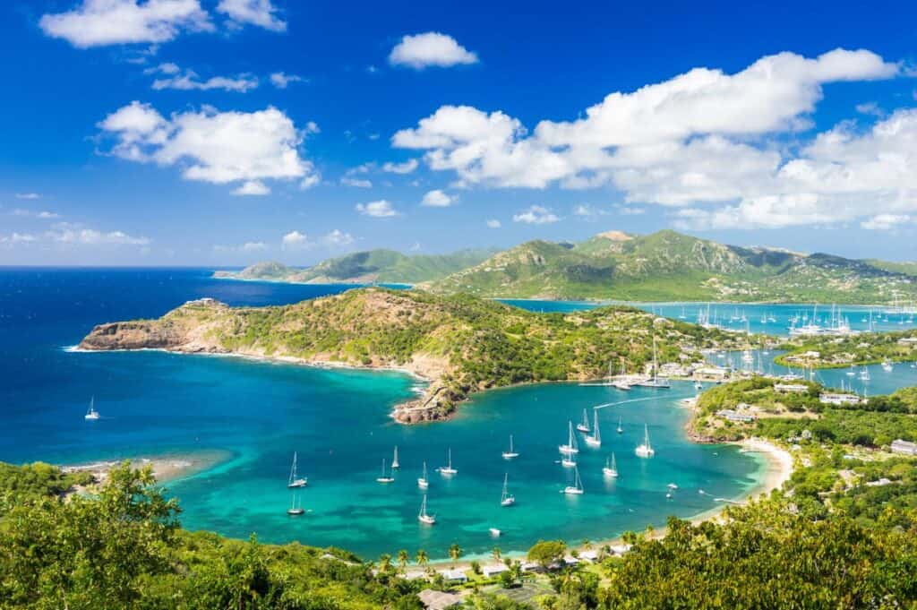 A scenic view of a Caribbean paradise with blue waters, green hills, and several sailboats anchored in the bay under a blue sky with scattered clouds.