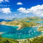 A scenic view of a Caribbean paradise with blue waters, green hills, and several sailboats anchored in the bay under a blue sky with scattered clouds.