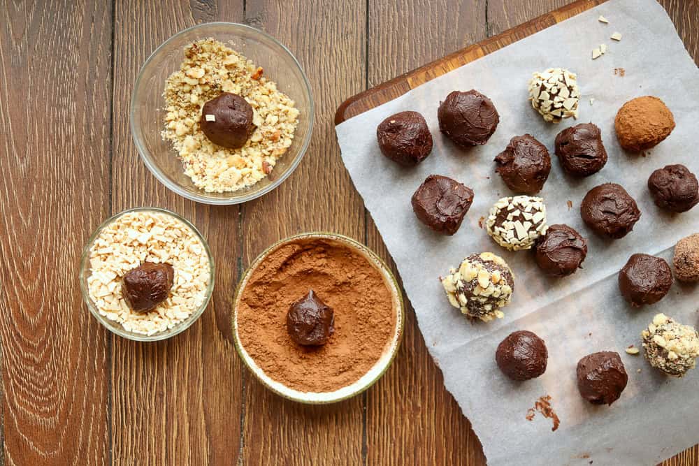 Several chocolate truffles are being coated in chopped nuts and cocoa powder on a wooden table. A plate with partially coated truffles and bowls with toppings are visible.