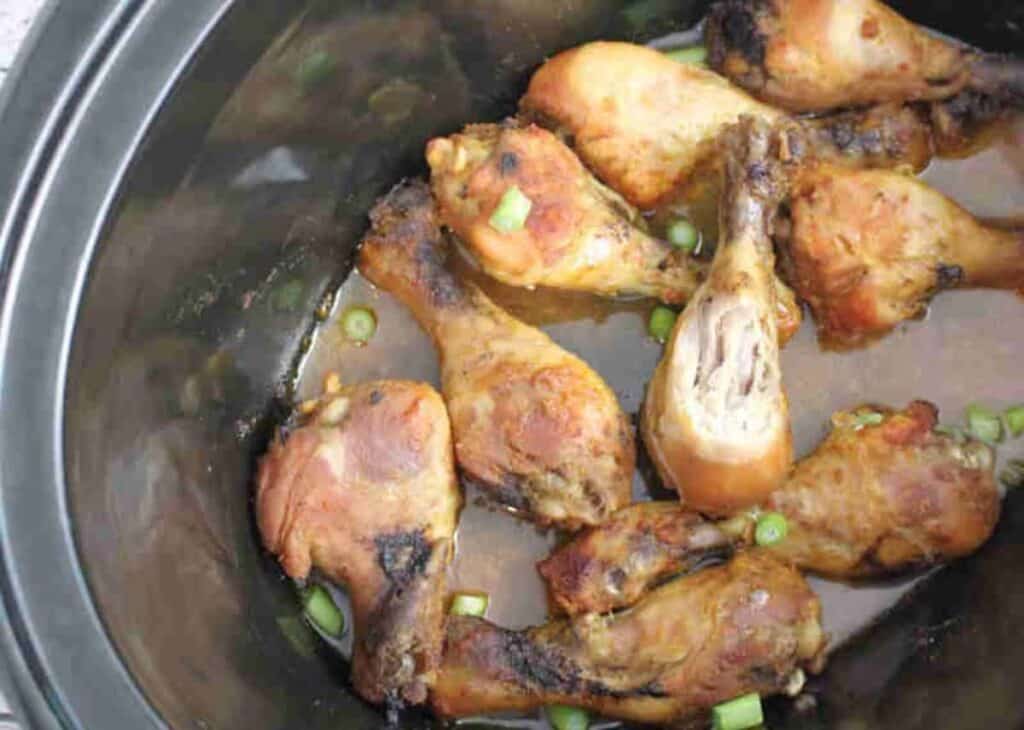 A slow cooker containing cooked chicken drumsticks in a brown sauce, garnished with chopped green onions.