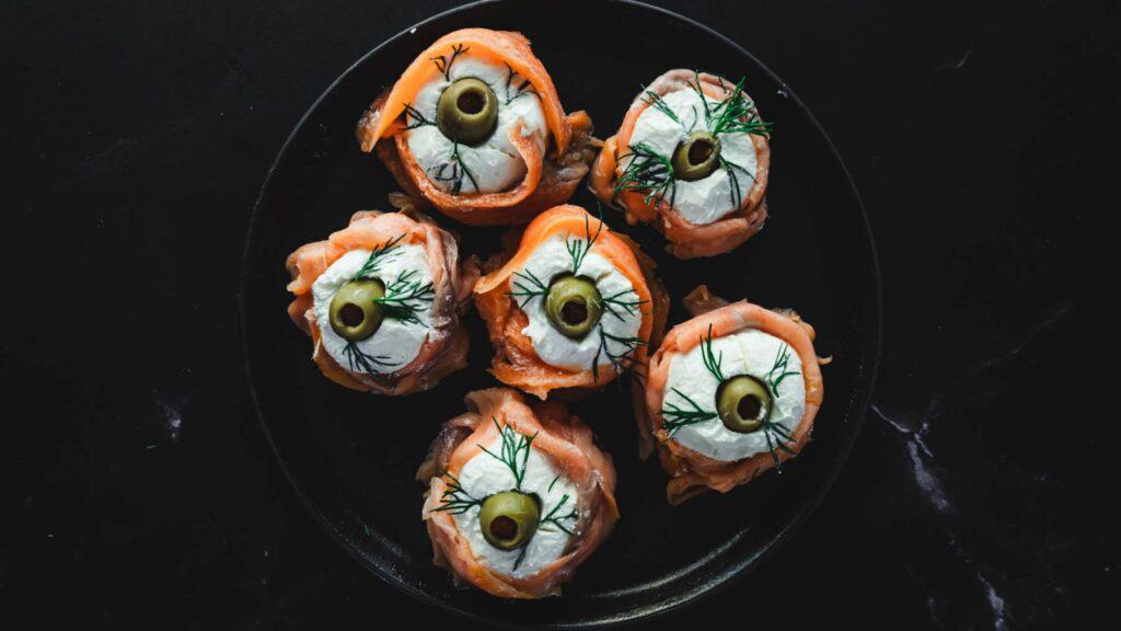 A black plate with six smoked salmon rolls filled with cream cheese, topped with green olives and sprigs of dill against a dark background.