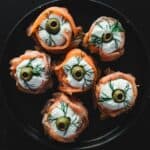 A black plate with six smoked salmon rolls filled with cream cheese, topped with green olives and sprigs of dill against a dark background.