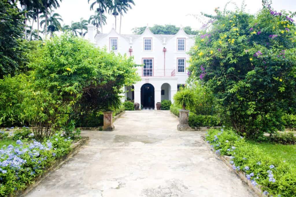 A colonial-style white mansion with arched entrances and red trim, surrounded by lush greenery and flowering plants, with a paved walkway leading to the front, evokes the rich history of rum in Barbados.