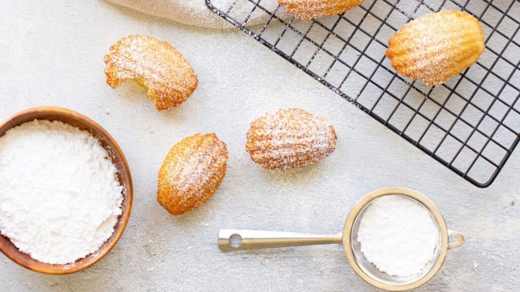 Madeleine cookies dusted with powdered sugar are on a cooling rack and scattered on a table next to a bowl and a sifter filled with more powdered sugar.