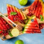 Grilled watermelon slices on a wooden board garnished with mint leaves, blueberries, red currants, and a honey dipper with lime halves on the side—truly one of those weird summer food combos that's surprisingly delightful.