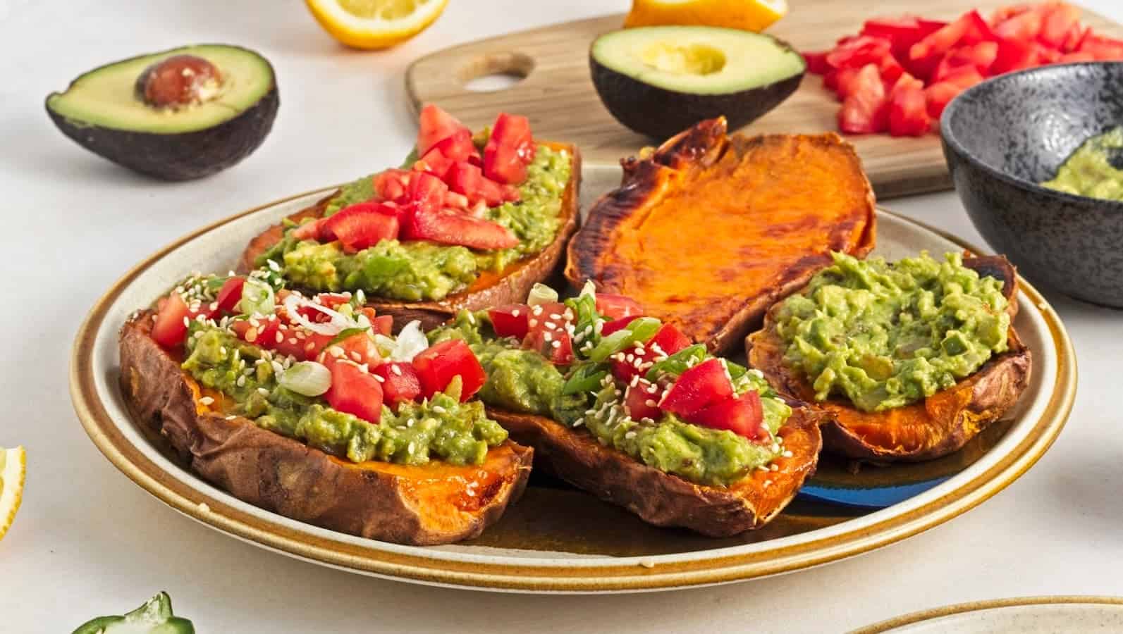 Plate of sweet potato halves topped with guacamole, chopped tomatoes, and sesame seeds. Halved avocado and chopped tomatoes in the background.
