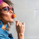 A person with curly hair, wearing red sunglasses, blue hoop earrings, a denim jacket, and a purple hairband, is sucking on a nostalgic candy lollipop.