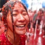 A person smiles while covered in tomato pulp during a crowded street festival.