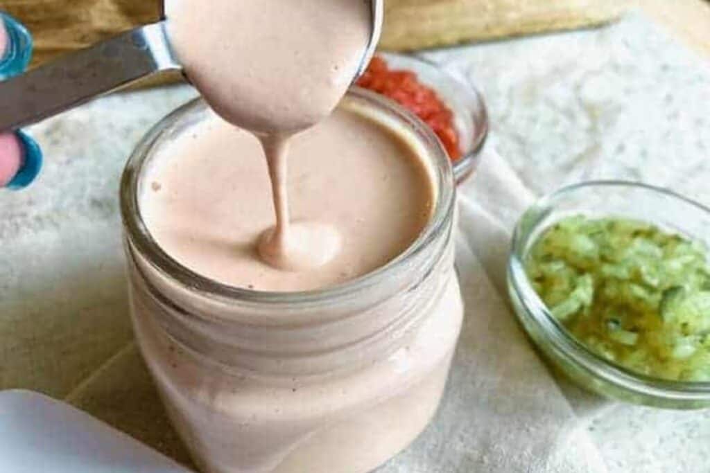 A spoon pours creamy sauce into a glass jar, surrounded by small bowls of diced tomatoes and chopped green onions on a table.