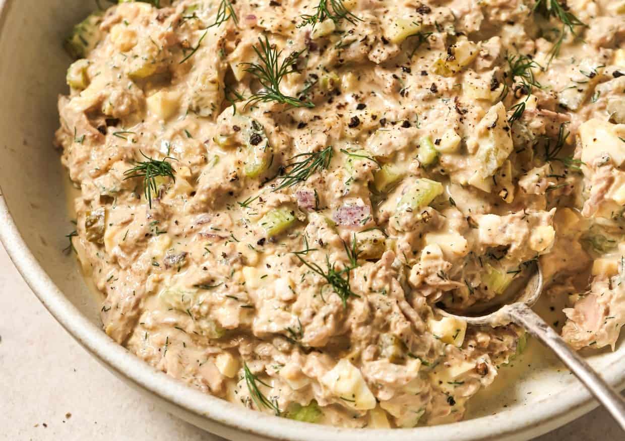 A close-up of a bowl of tuna egg salad mixed with celery, onions, and dill, topped with freshly ground black pepper, with a spoon placed on the side.