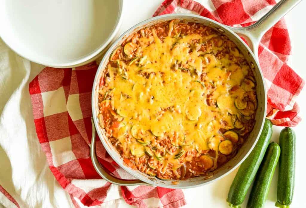 A skillet filled with a cheesy vegetable casserole, topped with melted cheese, next to a checkered red and white cloth and three whole zucchinis.